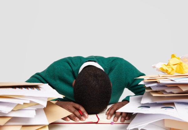 Stressful workaholic keeps head down on desk, feels tired and overworked, has much work, prepares for upcoming exam, writes information in diary, studies documentation, isolated on white wall